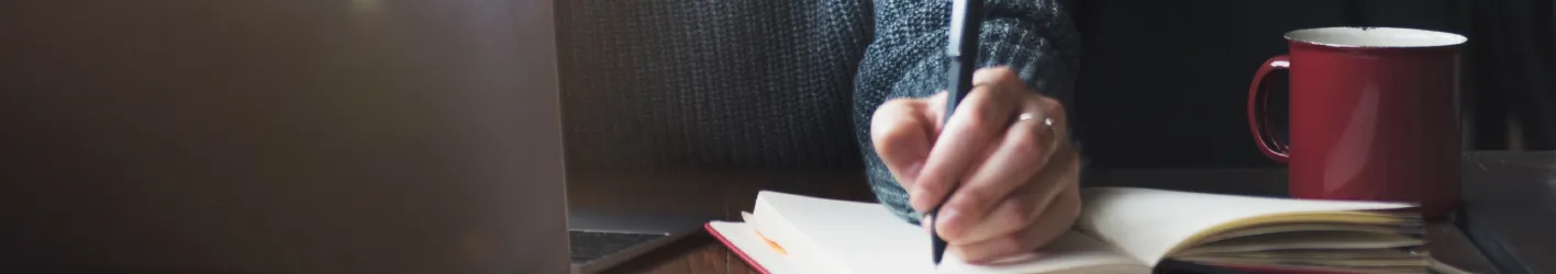 Person writing in a book with a red mug in the background.