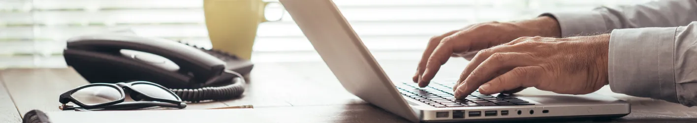 Employee typing on a laptop on a desk with a phone and glasses in the background